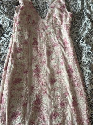 Rosa och vit spetsklänning med v-ringning - Söt klänning i vitt och rosa med blommigt spetsmönster och djup v-ringning. Klänningen är ärmlös och har en figurnära passform som framhäver siluetten. Perfekt för sommarens tillfällen och studentfirande.