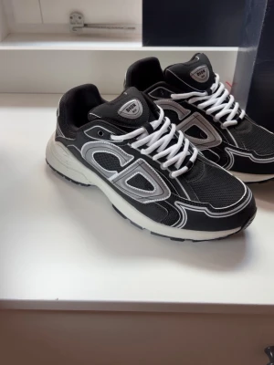 Svarta CD sneakers med vit sula - Snygga svarta CD sneakers med vit sula och vita detaljer. Skorna har en tydlig CD-logga på sidan, vita skosnören och meshpartier för extra komfort. Perfekt för dig som vill ha en trendig och exklusiv look. Materialet är en mix av textil och syntet.
