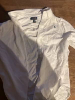 Vit skjorta från Polo Ralph Lauren - Klassisk vit skjorta från Polo Ralph Lauren med button-down krage och den ikoniska broderade loggan på bröstet. Skjortan har lång ärm och knappar framtill. Perfekt till en clean och stilren look.