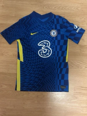 Chelsea FC matchtröja blå Nike - Chelsea FC matchtröja i blått med gula detaljer och grafiskt mönster. Tröjan har klubbmärke på bröstet, Nike-logga och sponsortryck framtill. Tillverkad i lätt och ventilerande material, perfekt för fotbollsträning eller match.