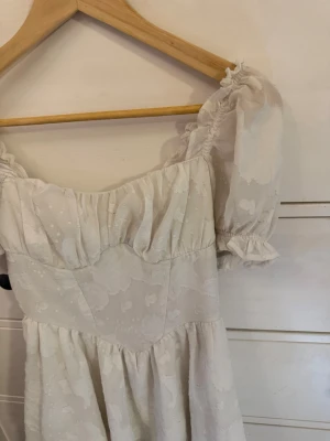 Vit blommig kortklänning med puffärm - Romantisk vit kortklänning med puffiga kortärmar och blommigt mönster. Klänningen har volangdetaljer och en lätt rynkad byst för en feminin look. Perfekt för vår och sommar, tillverkad i ett tunt och luftigt material.