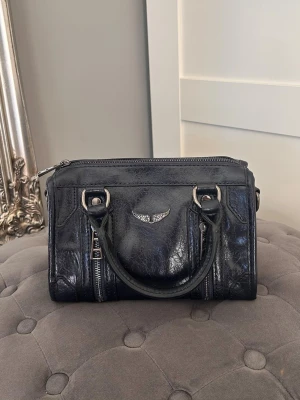 Snygg handväska  - Cool handväska. Väskan har dubbla handtag, silverfärgade detaljer, dragkedjor framtill och en ikonisk vinge i metall på framsidan. Perfekt storlek för det viktigaste och ger en edgy vibe till din outfit.