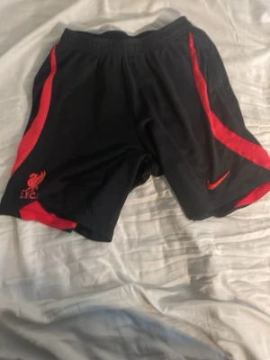 Liverpool FC träningsshorts Nike - Snygga svarta träningsshorts från Nike med röda detaljer och Liverpool FC-logga på ena benet. Shortsen har elastisk midja och är perfekta för fotboll eller träning. Tillverkade i lätt och bekvämt material.