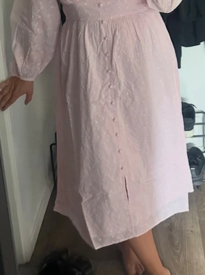 Ljusrosa långklänning med knappar - Långklänning i ljusrosa med diskret broderat mönster och knappar framtill. Klänningen har långärmade puffärmar och en slits nedtill. Perfekt för dig som gillar romantiska detaljer och en feminin look.