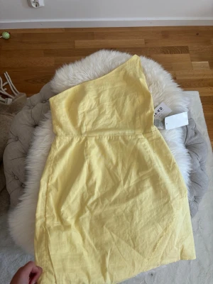 Gul oneshoulder kortklänning - Snygg gul kortklänning med oneshoulder-design och omlott framtill. Klänningen har en fräsch och somrig look, perfekt för varma dagar. Tillverkad i ett lätt material som ger en skön känsla och sitter snyggt på kroppen. 