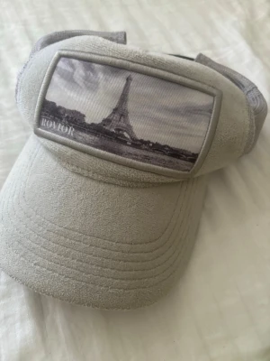 Beige keps med Eiffeltornet från Rovior - Snygg beige keps från Rovior med en stor patch framtill som visar ett svartvitt foto av Eiffeltornet. Kepsen har mesh på insidan och justerbar rem bak. Materialet är mjukt och mockaliknande på utsidan, perfekt för en trendig look.