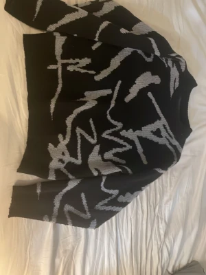 Svart stickad tröja med grått mönster - Cool svart stickad tröja med abstrakt grått mönster över hela plagget. Tröjan har långa ärmar och ribbstickade muddar vid nederkant och ärmslut. Perfekt för dig som gillar grafiska detaljer och vill ha något unikt i garderoben.  Passar någon som är cirka längden 168-175. 