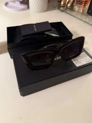 Svarta cat eye solglasögon från Prada - Snygga svarta solglasögon från Prada i trendig cat eye-modell. Glasögonen har mörka linser och en stilren, glansig båge med diskret Prada-logga på sidan. Perfekta för dig som vill ha en lyxig accessoar med attityd. Fint skick och kommer med låda och tillbehör, en repa som syns på sista bilden