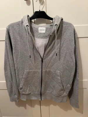 Grå zip hoodie från Jack & Jones - Mjuk grå hoodie från Jack & Jones med dragkedja framtill, två stora fickor och justerbar huva med snören. Insidan är fodrad med vitt fleece-tyg för extra komfort. Perfekt för chill dagar eller när du vill ha något bekvämt och stilrent.
