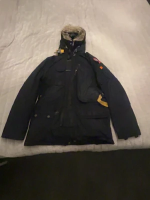 Mörkblå Parajumpers parkas med päls - Säljer en mörkblå parkas från Parajumpers med avtagbar huva och pälsdetalj. Jackan har flera fickor, gul detalj vid dragkedjan och klassisk Parajumpers-logga på ärmen. Perfekt för kalla dagar och riktigt snygg streetstil. Är i storlek xs men passar s