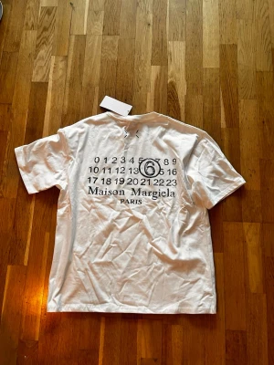 Maison Margiela vit t-shirt med tryck - Vit t-shirt från Maison Margiela med ikoniskt siffertryck och logga på ryggen samt diskreta stygn på bröstet. Klassisk passform och rund hals. Tillverkad i mjuk bomull som känns skön mot huden. Perfekt för dig som gillar minimalistisk och stilren design.