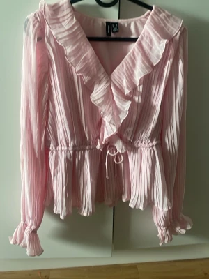Rosa blus med volang - Rosa blus från Vera Moda med volangkant längs v-ringningen och knyt i midjan. Blusen har långa ärmar med resår vid handlederna. Använd ett fåtal gånger