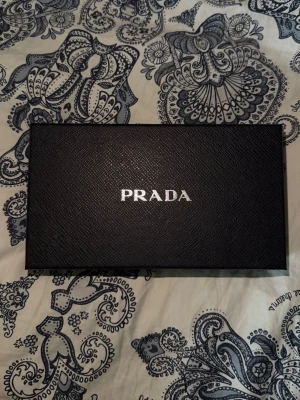 Svart plånbok från Prada - Snygg svart plånbok från Prada i exklusivt läder med klassisk guldfärgad logga framtill. Plånboken har en stilren och elegant design med rektangulär form och kommer i originalask. Perfekt för dig som vill ha en lyxig accessoar. Kvitto och certifikat finns, skriv dm för fler frågor eller bilder!