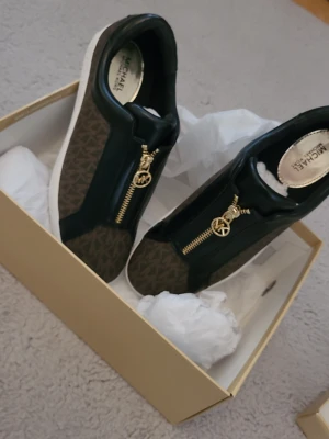 Michael Kors  - Snygga sneakers från Michael Kors i brunt och svart med guldfärgad dragkedja och MK-logga framtill. Skorna har ett mönstrat tyg med klassiskt MK-print och vita sulor. Materialmix av läder och textil ger en exklusiv känsla och modern look. Helt nya aldrig använda endast provade, karton och allt är med. Det är i storlek 38,5