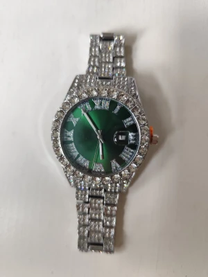 Grön glittrig klocka med stenar - Snygg klocka med grön urtavla och silverfärgat armband täckt av glittriga stenar. Klockan har romerska siffror och datumvisning. Perfekt för dig som gillar att sticka ut med accessoarer som verkligen syns. Minutvisaren är lite lös