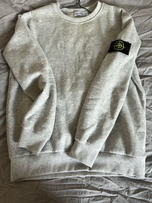 Grå sweatshirt från Stone Island - Säljer en stilren grå sweatshirt från Stone Island med klassisk rund hals och ribbade muddar. Tröjan har den ikoniska svarta patchen med logga på vänster ärm. Perfekt för dig som gillar streetwear och enkel design.