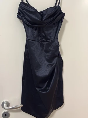 Mörkblå satin klänning med smala band - Snygg mörkblå klänning i satin med smala axelband och draperad urringning. Klänningen har en asymmetrisk nederkant och figurnära passform som ger en elegant siluett. Perfekt för dig som vill ha en stilren och trendig look. Klänningen är ifrån Black Silver ifrån Göteborg och ordinarie priset är 599kronor. Klänningen är i one size. Men den passar mig som har M.