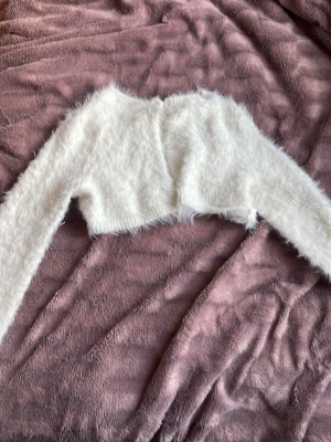 Vit fluffig bolero från H&M - Supermjuk och fluffig vit bolero från H&M. Kort modell med rund halsringning och långhårig, lurvig textur som ger en mysig känsla. Perfekt att slänga över en topp för en extra touch. Stängs framtill med en liten knapp.