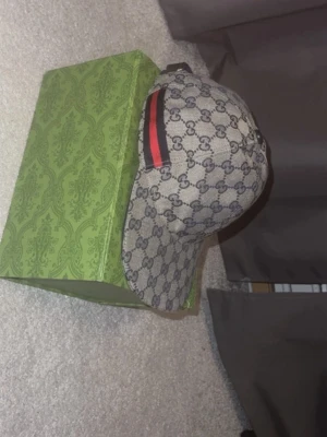 Gucci keps med GG-mönster - Säljer en snygg keps från Gucci med klassiskt GG-mönster i beige och mörkblått. Kepsen har en böjd skärm och detaljer med röd och svart rand på sidan. Materialet är canvas och kepsen har justerbar rem baktill.Priset kan diskuteras och det är inte hugget i sten.
