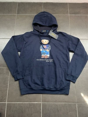 Mörkblå hoodie Polo Bear Ralph Lauren - Mörkblå hoodie från Ralph Lauren med den ikoniska Polo Bear-trycket på bröstet. Hoodien har huva med dragsko, magficka och ribbade muddar. Tillverkad i mjukt bomullsmaterial och har en avslappnad passform. Perfekt för dig som gillar streetwear och klassiska märken. Dessutom är den ganska tunn så den är perfekt för de kommande sommar kvällarna, passar även S