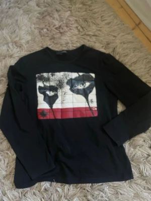 Svart långärmad topp från Dsquared2 - Svart långärmad topp från Dsquared2 med tryck framtill i vitt, rött och svart. Toppen har en klassisk rund halsringning och är tillverkad i mjuk bomull. Dsquared2-logga syns även i nacken. Perfekt för en cool och avslappnad stil.