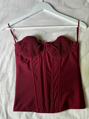 Vinröd korsett (strapless)  - Snygg vinröd korsett med smockade kupor. Framsidan har dekorativa sömmar och ett litet band mellan kuporna. Materialet är mjukt och stretchigt, perfekt för att skapa en cool och feminin look. (Banden är inte en del av looken!!)