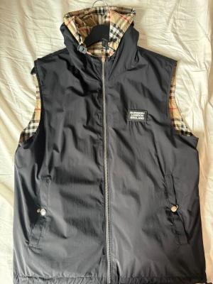 Svart dunväst från Burberry - Stilren svart dunväst från Burberry med klassiskt rutigt foder i beige, svart och rött. Västen har huva, dragkedja framtill, två fickor med tryckknappar och en patch med Burberry London England på bröstet. Du kan även vända den så att du har det klassiska Burberry mönstret. Storlek är S men står L för köptes i Dubai där storleken är inte samma.