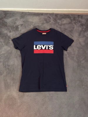 Svart Levi's t-shirt med logga - Snygg svart t-shirt från Levi's med stor logga i vitt, blått och rött på bröstet. Klassisk passform och rund halsringning med röd detalj på insidan. Perfekt till jeans eller shorts för en avslappnad look.
