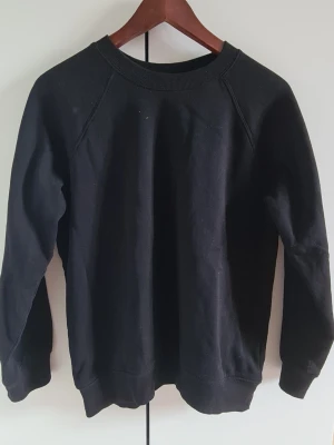 Svart basic sweatshirt - En enkel svart sweatshirt med rund halsringning och ribbade muddar vid ärmslut och nederkant. Tröjan har en avslappnad passform och är tillverkad i mjukt bomullsmaterial. Perfekt att matcha med jeans eller joggers.
