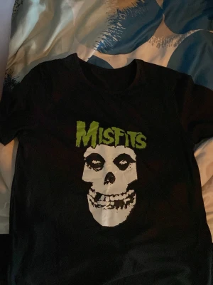Svart Misfits t-shirt med tryck - Säljer en svart t-shirt med ikoniskt Misfits-tryck i vitt och grönt framtill. T-shirten har klassisk passform och korta ärmar, perfekt för dig som gillar bandmerch och vill sticka ut med en cool look.