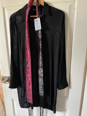Svart skjorta från Desigual med mönstrad scarf - Säljer en svart långärmad skjorta från Desigual i storlek 42. Skjortan har subtilt mönster i tyget och medföljer en färgglad scarf i rött och svart med vita detaljer och fransar. Klassisk krage och knappdetalj vid halsen. Perfekt för dig som gillar unika detaljer.