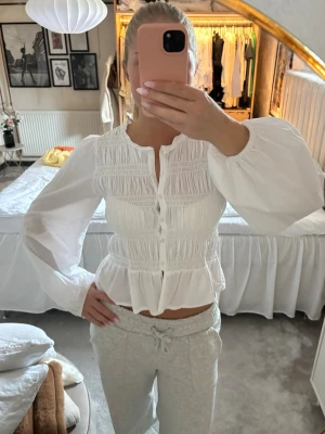 Vit blus med puffärm och smock - Säljer en vit blus med långa puffärmar och smockdetaljer över bröstet. Blusen har knappar framtill, rundad halsringning och volang nedtill. Tillverkad i ett lätt och luftigt tyg som ger en romantisk vibe. Använder inte den så mycket längre så därav säljer jag den🤍