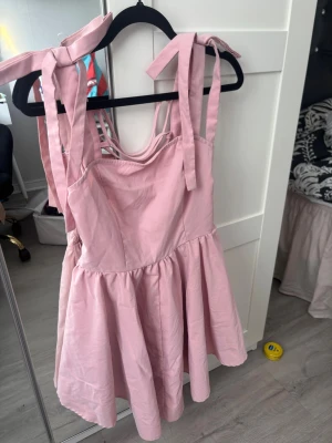 Rosa kort klänning med knytband - Supersöt ljusrosa kort klänning med breda axelband som knyts upptill och smock i ryggen. Klänningen har en volangig kjol och omlott-detaljer i ryggen. Perfekt för sommarens alla tillfällen!