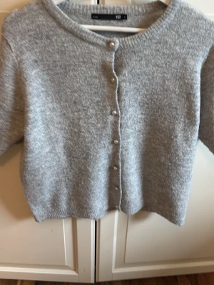 Grå kofta från Gina Tricot - Mysig gråmelerad kofta från Gina Tricot med rund halsringning och knappar framtill. Koftan har en rak passform, ribbade muddar och är tillverkad i mjukt stickat material. Perfekt att slänga över en topp för en enkel och stilren look.