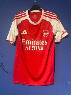 Arsenal Hemmatröja – Gyökeres 14 – Storlek S - Säljer en snygg Arsenal hemmatröja i storlek S med Gyökeres 14 tryckt på ryggen.  ✅ Storlek: S ✅ Färg: Röd/Vit ✅ Sponsor: Emirates Fly Better ✅ Spelartryck: Gyökeres 14 ✅ Skick: Mycket fint skick (se bilder)  Perfekt för Arsenal-supportrar eller samlare som vill ha en unik tröja med Gyökeres på ryggen.