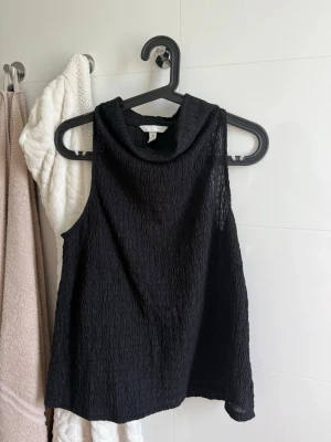 Svart ärmlös topp med struktur - Säljer en svart ärmlös topp från H&M med hög krage och snyggt strukturerat tyg. Toppen har en loose passform och är perfekt för dig som gillar stilrena plagg med det lilla extra. Passar till många olika looks och är superenkel att styla.