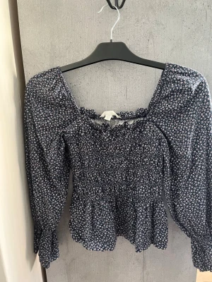 Blommig blå blus från H&M - Supersöt blus från H&M i mörkblått med små vita och röda blommor. Blusen har smock över bröstet, volang nertill och en fyrkantig ringning. Långärmad modell med puffiga ärmar och elastiska muddar. Perfekt för att skapa en trendig look.