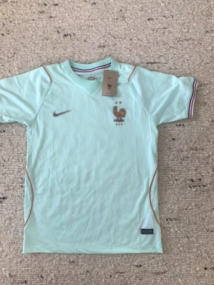 Ljusblå Frankrike Nike bortatröja VM 2026 - Snygg ljusblå matchtröja från Nike med Frankrikes landslagsemblem och två stjärnor broderade på bröstet. Tröjan har korta ärmar, V-ringning och tunna orange detaljer längs sidorna. Kragen har en diskret trikolor-detalj på insidan.