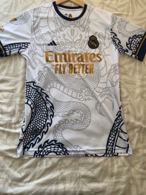 Real Madrid draktröja Ronaldo 7 - Snygg Real Madrid matchtröja från Adidas med Ronaldo 7 på ryggen. Tröjan är vit med grått och svart drakmönster, guldiga detaljer och klubbmärke på bröstet. Tillverkad i lätt och ventilerande material, perfekt för fotboll eller streetwear. Den är size s men passar en man på 185 cm 