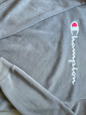 Grå sweatshirt från Champion - Snygg grå sweatshirt från Champion med broderad logga i vitt och rött på bröstet. Tröjan har rund halsringning, långa ärmar och är tillverkad i mjuk bomullsblandning. Perfekt för en avslappnad och sportig stil.