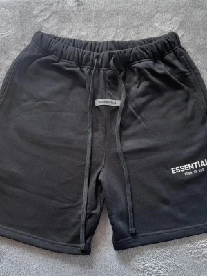 Svarta shorts från Essentials Fear of God - Snygga svarta shorts från Essentials Fear of God med vit logga på vänster ben. Shortsen har elastisk midja med snörning och är tillverkade i mjukt material som passar perfekt till sommaren. Klassisk och clean design med normal passform.