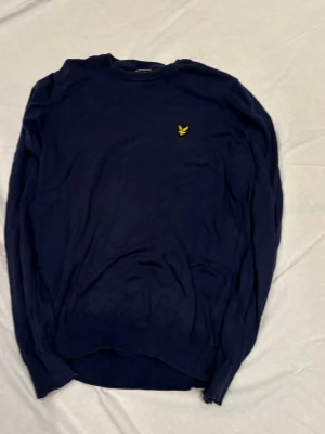 Mörkblå stickad tröja Lyle & Scott - Mörkblå stickad tröja från Lyle & Scott med rund halsringning och den klassiska gula logon broderad på bröstet. Tröjan har långa ärmar och ribbade muddar vid ärmslut och nederkant. Perfekt för lager på lager och enkel att matcha.