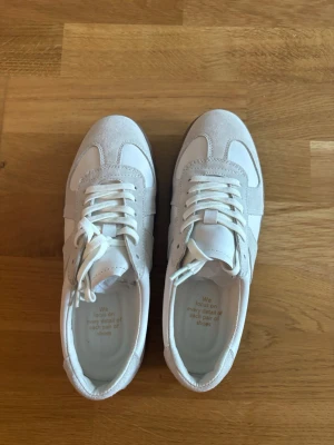 Vita sneakers med mockadetaljer/ Maison margiela - Stilrena vita sneakers med ljusgrå mockadetaljer och vita skosnören. Skorna har en klassisk låg siluett och en gummisula i grått. Materialmix av läder och mocka ger en snygg kontrast. Perfekta för dig som gillar minimalistisk och clean stil. Inspirerade av maison margiela sneakers. Strlk 38.5 men passar 39 också!