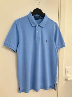 Ljusblå pikétröja från Ralph Lauren (M) - Klassisk ljusblå pikétröja från Ralph Lauren med korta ärmar och krage. Tröjan har två knappar framtill och den ikoniska broderade loggan på bröstet. Tillverkad i mjuk bomull för en bekväm känsla.