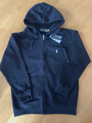 Svart hoodie från Polo Ralph Lauren - Snygg svart hoodie från Polo Ralph Lauren med dragkedja framtill och klassisk huva med dragsko. Ikoniska vita loggan broderad på bröstet. Tillverkad i mjukt bomullsmaterial och har ribbade muddar vid ärmar och nederkant. Perfekt för en avslappnad och stilren look.