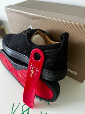 Svarta sneakers från CL - Snygga svarta sneakers från CL i mocka med klassisk röd sula och diskret logga på hälen. Skorna har svarta snören och detaljer i svart läder. Perfekta för dig som vill ha en stilren och exklusiv look.