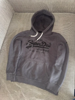 Mörkgrå Superdry hoodie med tryck - Säljer en mörkgrå hoodie från Superdry med svart logotyp och japansk text på bröstet. Hoodien har en stor ficka framtill, vita dragsnören i huvan och ribbade muddar vid ärmar och nederkant. Perfekt för en avslappnad streetwear-look.