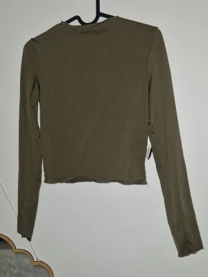 långärmad topp  - Säljer en olivgrön långärmad topp från Gina Tricot. Toppen är croppad med råa kanter och har en enkel, stilren design. Perfekt att matcha med jeans eller kjol för en cool look.
