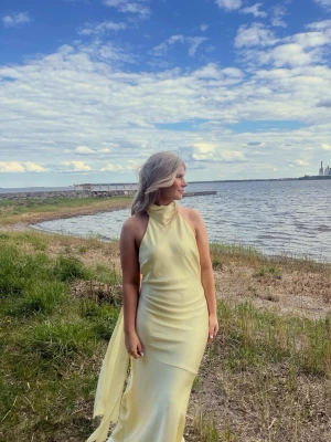 Ljusgul långklänning i satin - Elegant ljusgul långklänning i satin med halterneck och svepande siluett. Klänningen har en mjuk glans, figurnära passform och öppen i ryggen. Slutsåld på Gina Tricot