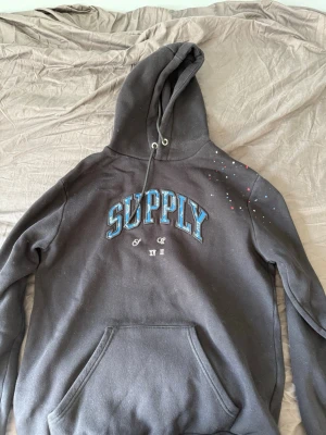 Svart track suit med blå SUPPLY-tryck - Svart tracksuit med stor känguruficka och huva med dragsko. Framsidan har ett blått SUPPLY-tryck och små vita detaljer. På ena axeln finns färgstänk i rött och blått. Tillverkad i mjukt bomullsmaterial, perfekt för en avslappnad stil. Både byxorna och hoodien säljs tillsammans som ett sammanhängande sett. Det går även att köpa endast hoodien eller byxorna men helst båda tillsammans 